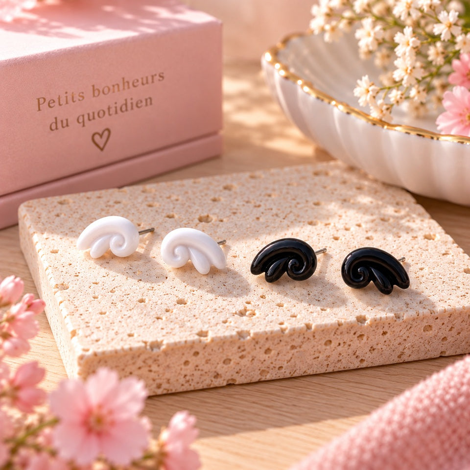 Deux paires de puces d’oreilles noires et blanches au design tourbillon, présentées sur un support en pierre dans un décor féminin rose et fleuri.
