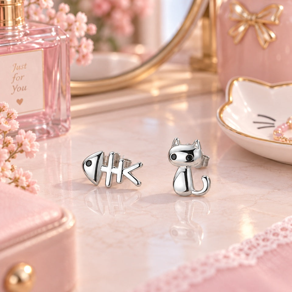 Puces d’oreilles asymétriques argentées en forme de chat et d’arête de poisson, posées sur une coiffeuse rose élégante dans un décor fleuri et féminin.