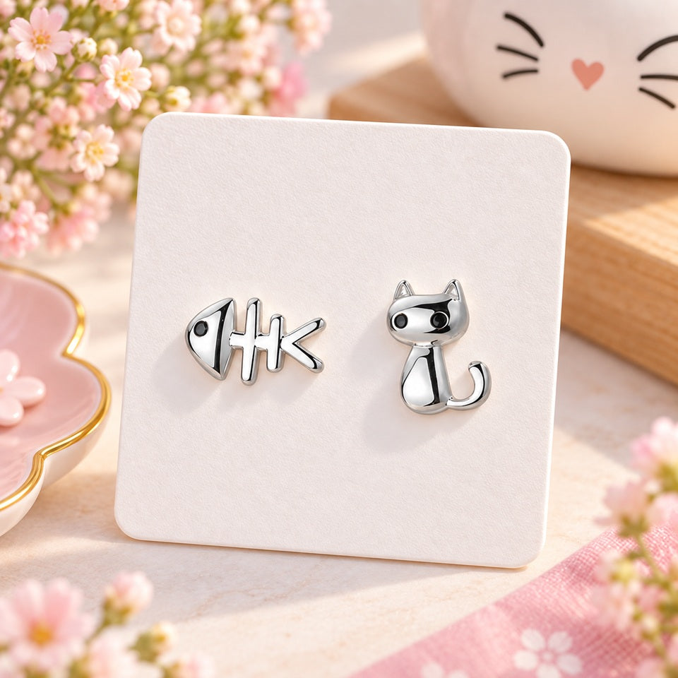 Puces d’oreilles asymétriques argentées en forme de chat et d’arête de poisson, présentées sur un support blanc dans un décor fleuri rose et kawaii.