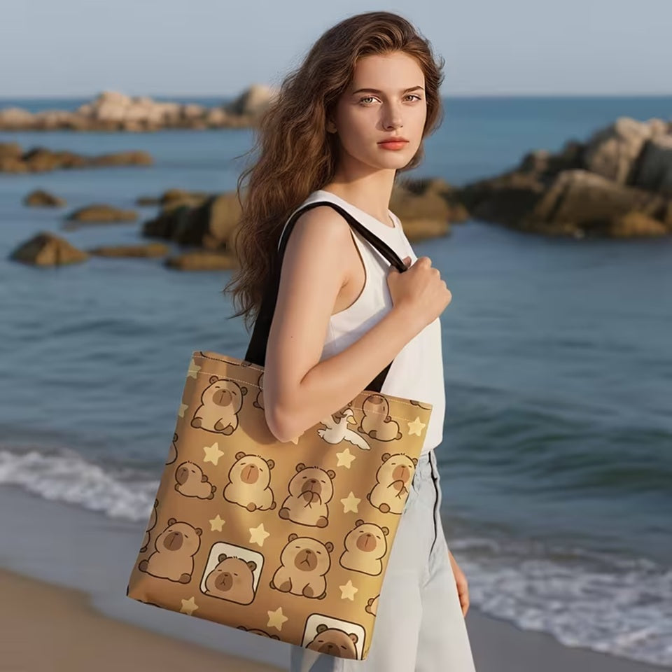 un sac fourre-tout marron au design de capybara et d'étoiles trop mignons. Il est porté à l'épaule par une femme habillé en blanc, au bord de la mer.