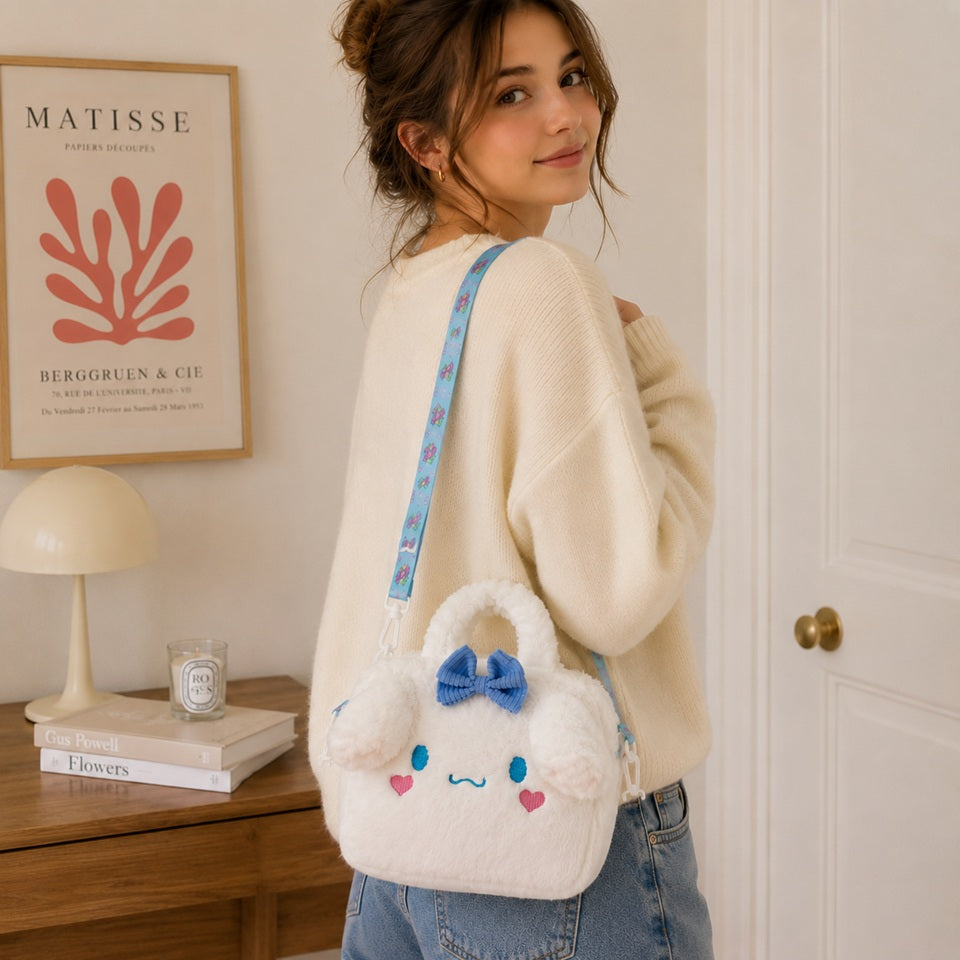 une petite sacoche toute douce et fluffy de Cinnamoroll. Elle est blanche et les yeux et des coeurs roses sont brodés dessus et arbore un joli noeud bleu. Elle est portée à l'épaule par une jeune femme.