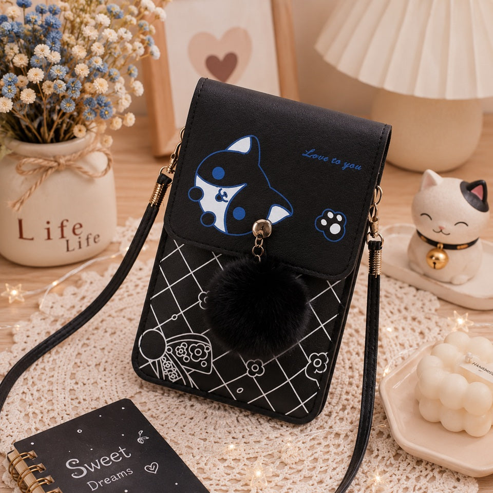 Sacoche téléphone noire kawaii avec motif chat, pompon noir et bandoulière, présentée dans un décor chaleureux et élégant avec fleurs séchées, lampe, figurine de chat, bougie et accessoires déco. Petit sac à bandoulière pour femme, pratique, compact et tendance.
