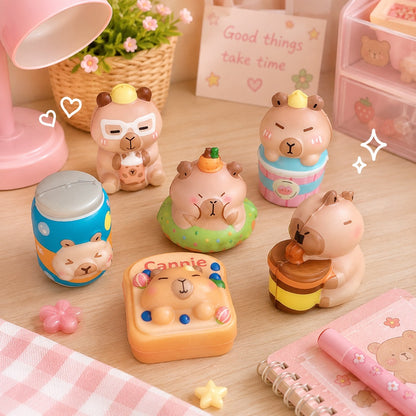 Une collection de squishies capybara aux couleurs pastel, disposés sur une jolie table en bois clair. Les figurines représentent des capybaras dans différents costumes et gourmandises (cupcake, boisson pain de mie, bubble tea), entourés d'une décoration douce avec une lampe rose, une plante, un carnet et de petits éléments décoratifs. L'ambiance est chaleureuse, lumineuse et mignonne.