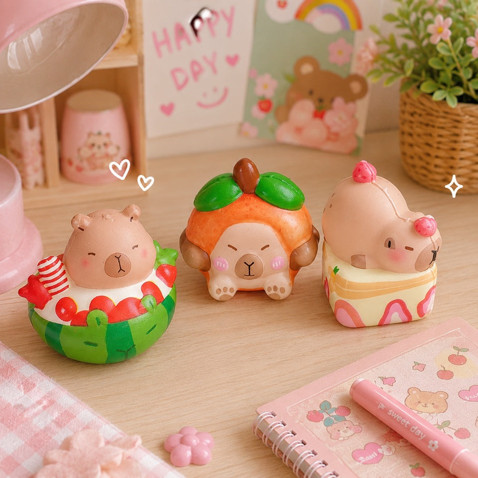 Trois squishies capybara kawaii sont présentés sur une jolie table en bois clair dans un décor pastel. Le premier flotte dans une pastèque, le deuxième porte un costume d'orange, et le troisième est allongé dans un petit dessert crémeux. En arrière-plan, une lampe rose, une plante, un carnet et de petits éléments décoratifs créent une ambiance douce, chaleureuse et gourmande, idéale pour mettre en valeur ces adorables jouets anti-stress.