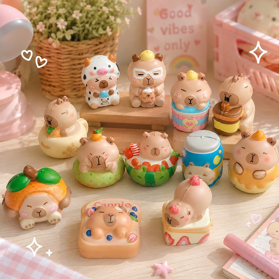Mockup kawaii présentant une collection de squishies capybara aux couleurs pastel, disposés sur une jolie table en bois clair. Les figurines représentent des capybaras dans différents costumes et gourmandises (lait, cupcake, miel, pastèque, agrumes, bain, etc.), entourés d'une décoration douce avec une lampe rose, une plante, un carnet et de petits éléments décoratifs. L'ambiance est chaleureuse, lumineuse et mignonne, idéale pour mettre en valeur ces jouets anti-stress.