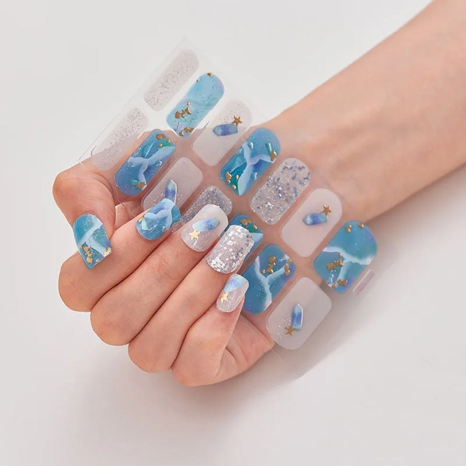 Stickers nail art bleu et blanc à paillettes avec motifs marins et détails dorés, présentés dans une main dont les ongles sont habillés avec les stickers.