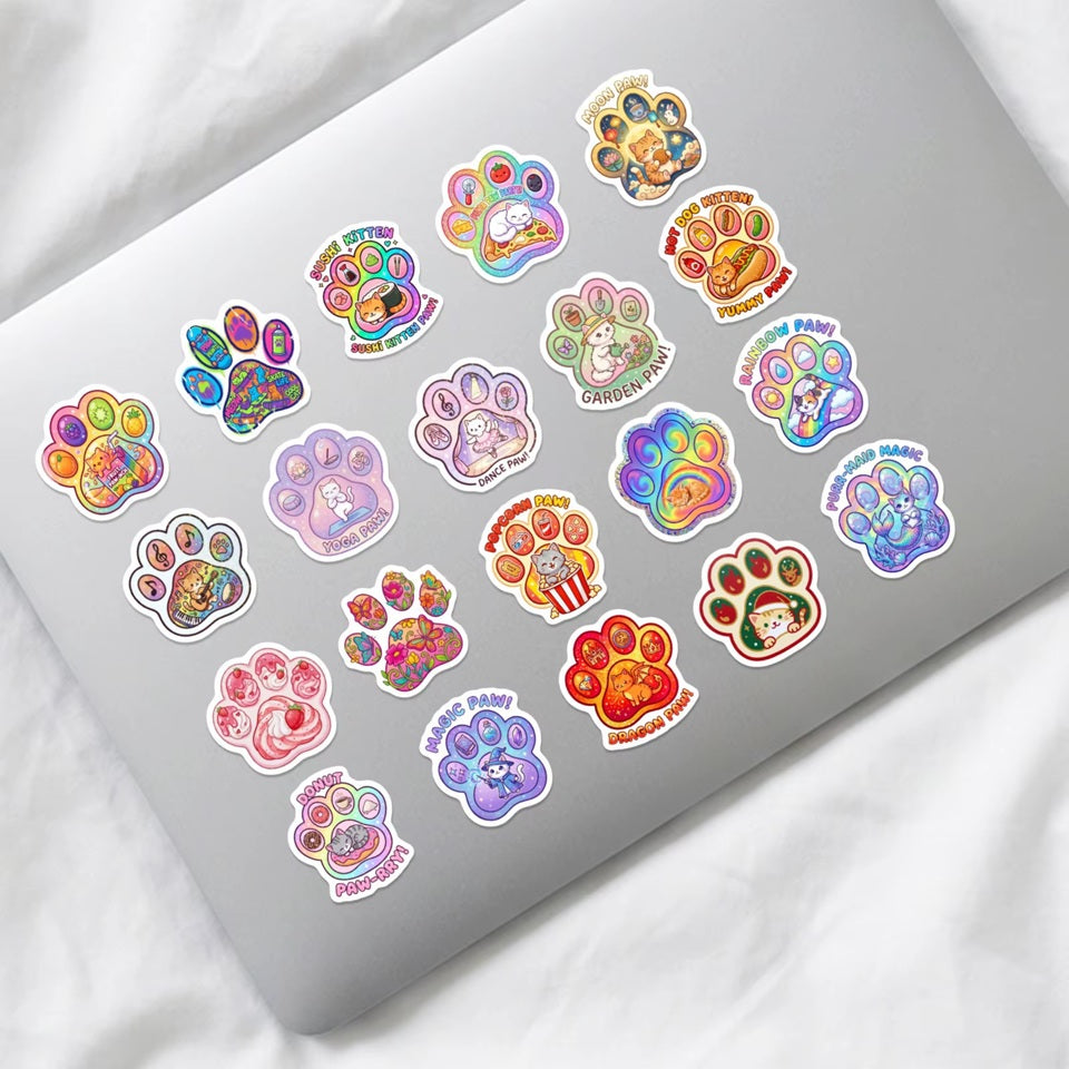 Gros plan sur des stickers en forme de pattes de chat colorées dans lesquelles se trouvent des scénettes adorables de chat dans des situations de la vie quotidienne : danse, musique, au bureau etc... Ils sont collés sur un ordinateur portable. Parfaits pour décorer d'une façon mignonne des accessoires.
