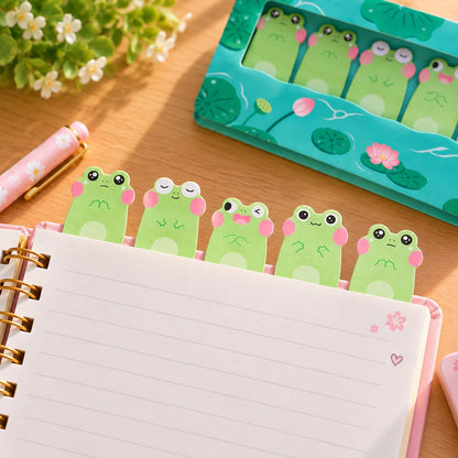 Lot de 5 mémos adhésifs kawaii en forme de grenouilles vertes aux joues roses et expressions rigolotes, collés sur le haut d’une page de cahier à spirales, avec leur étui turquoise décoré de nénuphars et de fleurs de lotus.
