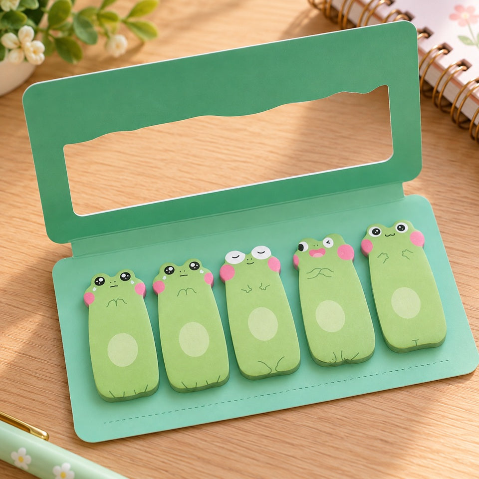 Lot de 5 blocs mémo adhésifs kawaii en forme de grenouilles vertes aux expressions différentes, présenté dans un étui turquoise ouvert sur un bureau en bois clair, avec carnet fleuri et accessoires de papeterie.