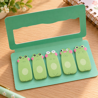 Lot de 5 blocs mémo adhésifs kawaii en forme de grenouilles vertes aux expressions différentes, présenté dans un étui turquoise ouvert sur un bureau en bois clair, avec carnet fleuri et accessoires de papeterie.
