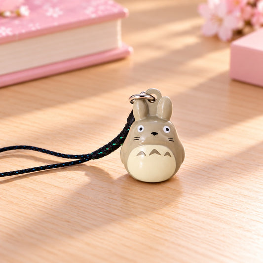 Porte-clés Totoro kawaii inspiré de l’univers du Studio Ghibli, représentant l’adorable personnage gris aux petites oreilles, au ventre crème et aux yeux expressifs. Cette figurine miniature est accompagnée d’une dragonne noire aux reflets bleutés et d’un anneau métallique argenté. Présenté sur une jolie table en bois clair, dans un décor pastel avec un carnet rose et quelques fleurs de sakura, cet accessoire japonais mignon est idéal pour personnaliser ses clés, son sac ou son téléphone.