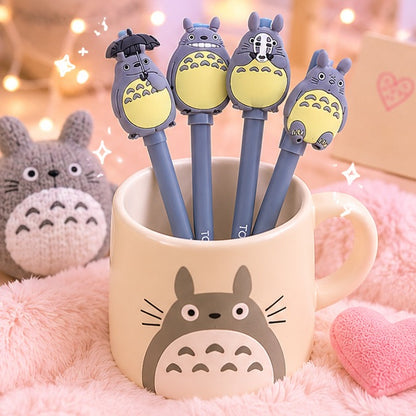 Stylos gel Totoro kawaii gris présentés dans une tasse Totoro beige, entourés d’une ambiance pastel chaleureuse avec peluche, coussin cœur rose et lumières douces en arrière-plan.