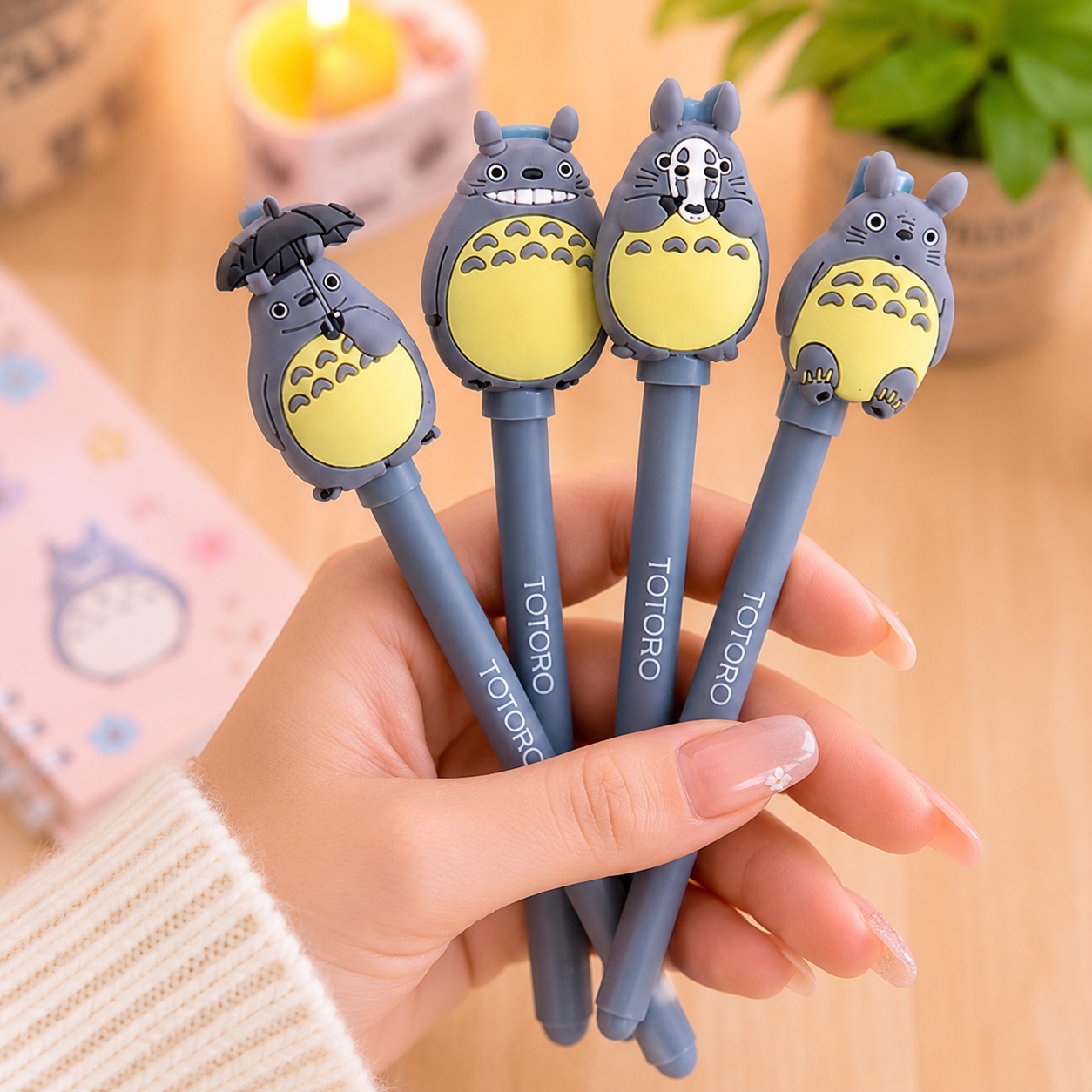 Main d’une femme manucurée tenant quatre stylos gel Totoro gris kawaii avec différents personnages inspirés de l’univers japonais, dans une ambiance pastel chaleureuse et mignonne.