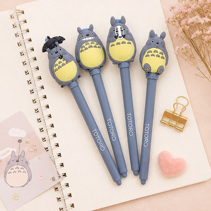 Quatre stylos gel Totoro gris kawaii posés sur un carnet beige, entourés d’accessoires pastel mignons comme un cœur en peluche rose, des fleurs séchées et une pince dorée dans une ambiance douce et cosy.