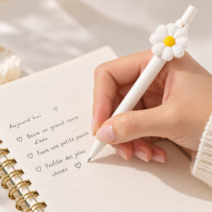 Main de femme écrivant dans un carnet avec un stylo blanc orné d’une marguerite, dans une ambiance douce, fleurie et lumineuse.