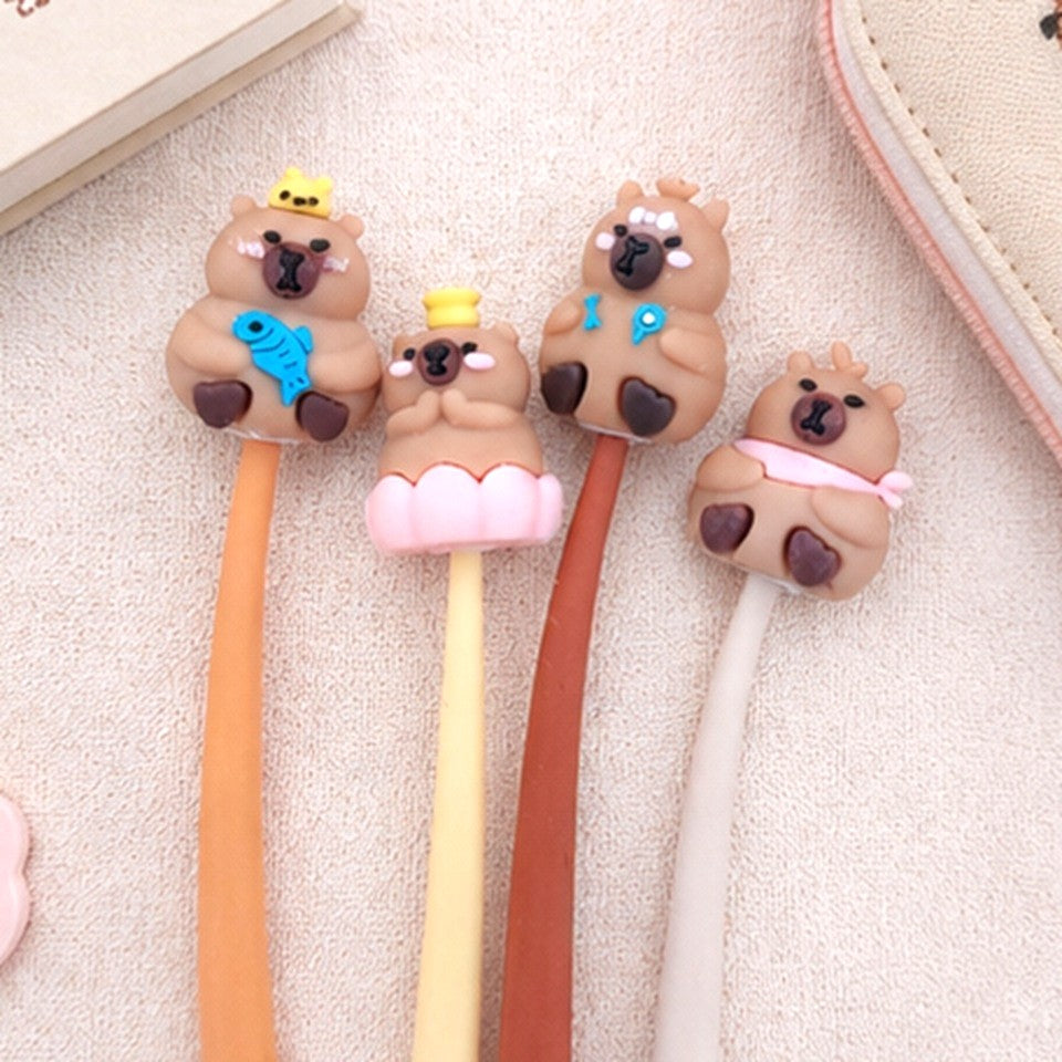 Quatre stylos kawaii souples ornés de petits capybaras adorables sont disposés côte à côte sur un tissu beige, à côté d'un carnet illustré et d'une trousse mignonne. Une mise en scène douce et chaleureuse met en valeur ces stylos fantaisie parfaits pour l'école, le bureau ou les amateurs de papeterie kawaii.