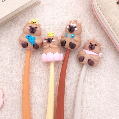 Quatre stylos kawaii souples ornés de petits capybaras adorables sont disposés côte à côte sur un tissu beige, à côté d'un carnet illustré et d'une trousse mignonne. Une mise en scène douce et chaleureuse met en valeur ces stylos fantaisie parfaits pour l'école, le bureau ou les amateurs de papeterie kawaii.