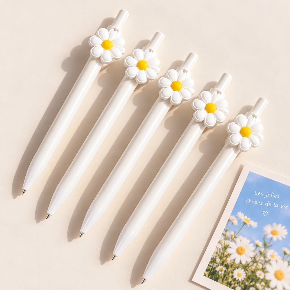 Stylos blancs kawaii ornés de marguerites blanches et jaunes, présentés avec un joli carnet fleuri dans une ambiance douce et estivale.