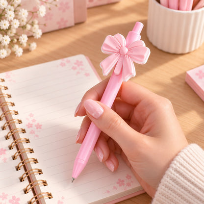 Stylo kawaii rose pastel orné d'un grand nœud rose en relief avec un délicat contour blanc, tenu dans la main manucurée d'une femme au-dessus d'un carnet ouvert décoré de fleurs de sakura. Présenté sur un bureau en bois clair avec des accessoires de papeterie rose, ce joli stylo au design chic, romantique et élégant est idéal pour écrire, décorer son bureau ou offrir un adorable cadeau aux amoureuses de papeterie kawaii.