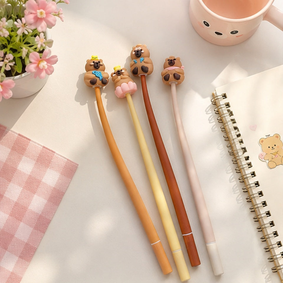 Stylos capybara souples kawaii disposés sur un carnet dans un joli décor pastel avec fleurs et tissu à carreaux roses. Lot de 4 stylos fantaisie mignons pour papeterie, école et bureau.