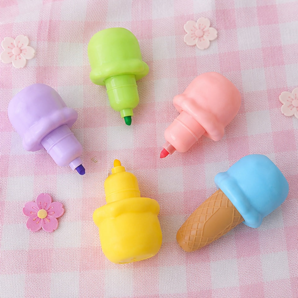 Dessurligneurs en forme de glaces colorées sont disposés sur une nappe à carreaux rose et blanc, accompagnés des différents embouts colorés représentant les boules de glace. Les capuchons vert, rose, violet, jaune et le cornet bleu sont présentés séparément pour mettre en valeur les différentes couleurs des surligneurs. De petites fleurs décoratives renforcent l'ambiance pastel, kawaii et gourmande de cette mise en scène.