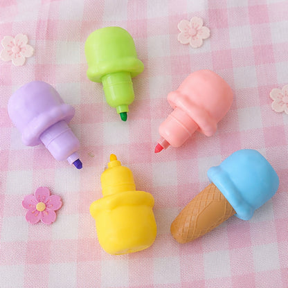 Dessurligneurs en forme de glaces colorées sont disposés sur une nappe à carreaux rose et blanc, accompagnés des différents embouts colorés représentant les boules de glace. Les capuchons vert, rose, violet, jaune et le cornet bleu sont présentés séparément pour mettre en valeur les différentes couleurs des surligneurs. De petites fleurs décoratives renforcent l'ambiance pastel, kawaii et gourmande de cette mise en scène.