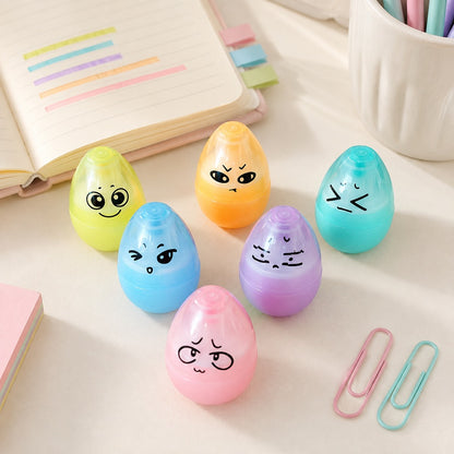 Un lot de six surligneurs en forme d’œufs kawaii aux couleurs pastel, présentés sur un bureau lumineux avec un carnet et des accessoires de papeterie.
