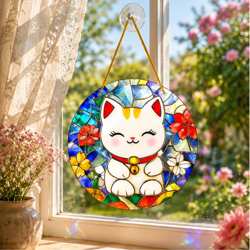 Suspension décorative ronde représentant un Maneki-neko en style vitrail, accrochée devant une fenêtre baignée de lumière naturelle. Les couleurs vives du verre teinté – bleu, vert, rouge, jaune et blanc – sont sublimées par les rayons du soleil. À l'arrière-plan, un jardin fleuri aux tons roses et verts apporte une ambiance douce et printanière, tandis qu'un vase de petites fleurs posé sur le rebord de la fenêtre complète cette mise en scène chaleureuse et apaisante.