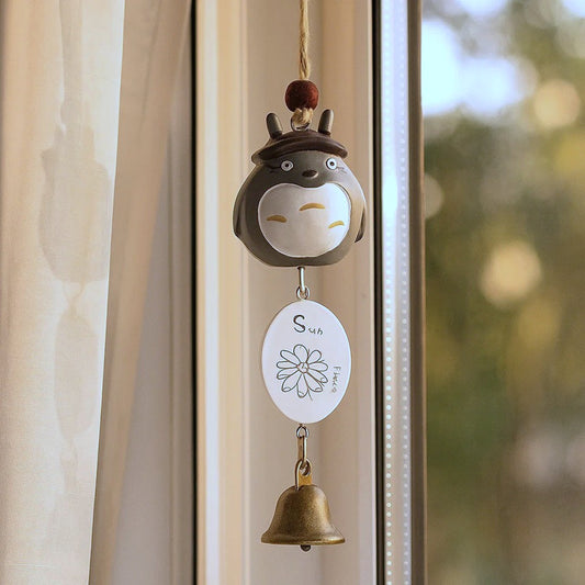 une suspension Totoro portant un petit chapeau de jardinier. une clochette et une joli epancarte en bois sont suspendus au bout. Il est très mignon et est à suspendre à un porche d'entrée ou une fenêtre