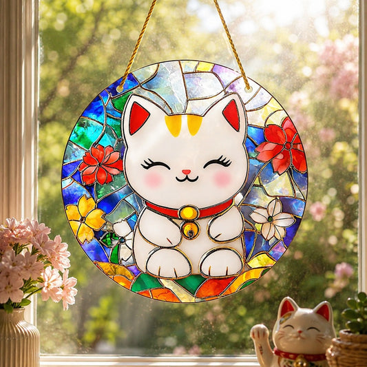 une suspension décorative en forme de vitrail représentant un Maneki-neko blanc souriant, orné d'un collier rouge et d'une clochette dorée. Suspendue devant une fenêtre ensoleillée par une cordelette jaune, la décoration laisse passer la lumière à travers ses motifs colorés bleu, vert, rouge, jaune et blanc, inspirés des fleurs et du verre teinté. En arrière-plan, un jardin verdoyant baigné de soleil ainsi que quelques éléments décoratifs créent une ambiance chaleureuse, lumineuse et apaisante.