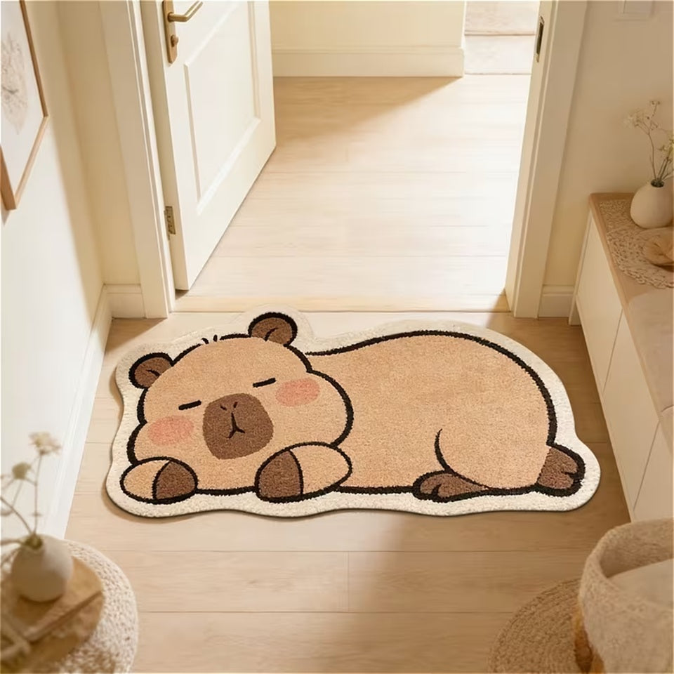Un tapis de sol en forme et aux couleurs d'un adorable capybara. Il est couché et semble dormir paisiblement. Il est posé au sol devant une porte, créant une ambiance douce et calme.