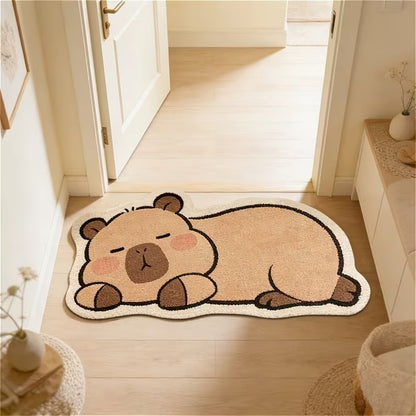 Un tapis de sol en forme et aux couleurs d'un adorable capybara. Il est couché et semble dormir paisiblement. Il est posé au sol devant une porte, créant une ambiance douce et calme.