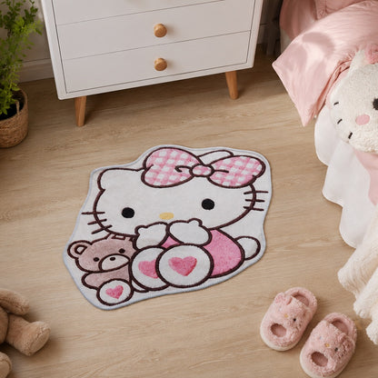 Tapis de sol en forme découpée représentant Hello Kitty tenant un ourson, dans des tons blanc, rose pastel et gris clair. Le tapis est posé sur un parquet clair dans une chambre d'enfant cosy, à côté d'un lit avec linge de lit rose, d'une commode blanche, de chaussons roses et de peluches. L'ensemble crée une ambiance douce, chaleureuse et kawaii.
