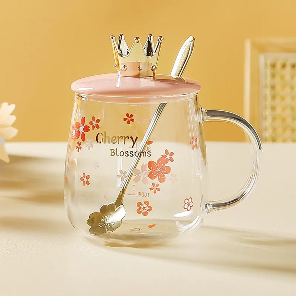Gros plan sur une tasse transparente au design de fleurs de sakura rose dont le couvercle rose est orné d'une couronne dorée. Elle a une cuillère en forme de fleur de sakura. Design floral mignon et coloré qui rappelle le Japon