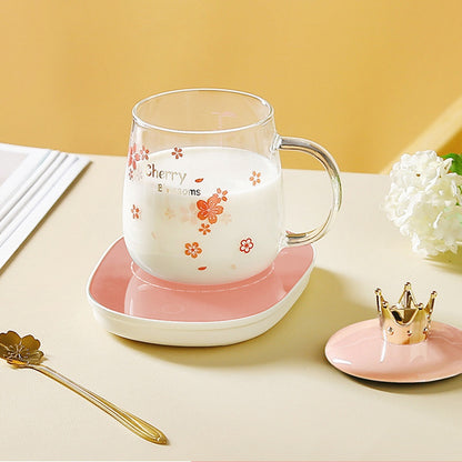 une tasse transparente au design de fleurs de sakura rose posée sur un support rose sur une table blanche. A côté, se trouve le couvercle rose orné d'une couronne dorée et la cuillère en forme de fleur de sakura. Design floral mignon et coloré qui rappelle le Japon