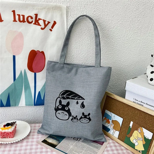 un joli tote bag gris illustré de Totoro et de ses amis en version chibi. Il porte une feuille au-dessus de lui.