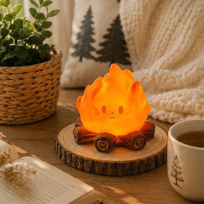Veilleuse feu de camp kawaii orange allumée sur socle effet bois, dans un décor cosy et chaleureux avec livre, tasse et plante verte.
