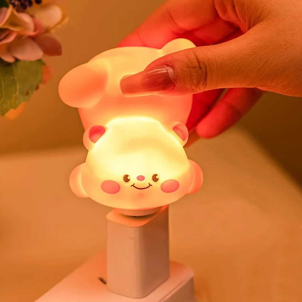 Gros plan sur une adorable veilleuse en forme d'ourson qui fait une galipette. Elle est branchée sur une prise USB dans une ambiance feutrée, douce et calme. Elle est tenue par une main de femme;