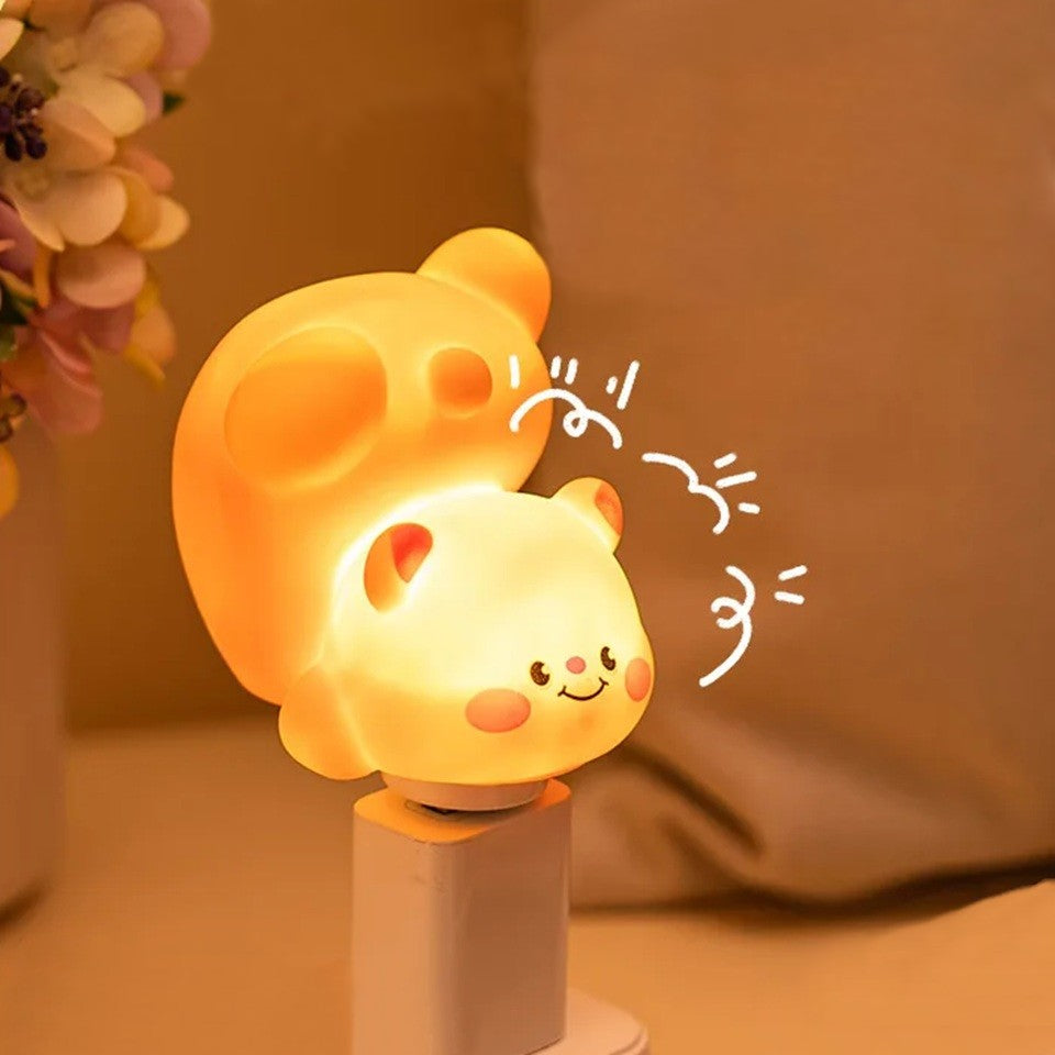 Gros plan sur une adorable veilleuse en forme d'ourson qui fait une galipette. Elle brille d'un ton chaud. Elle est branchée sur une prise USB dans une ambiance feutrée, douce et calme.